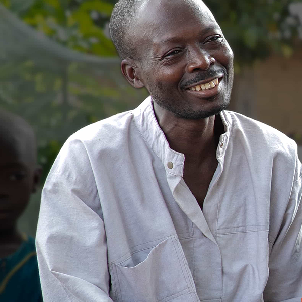 Man in a light gray shirt smiling outdoors with blurred background