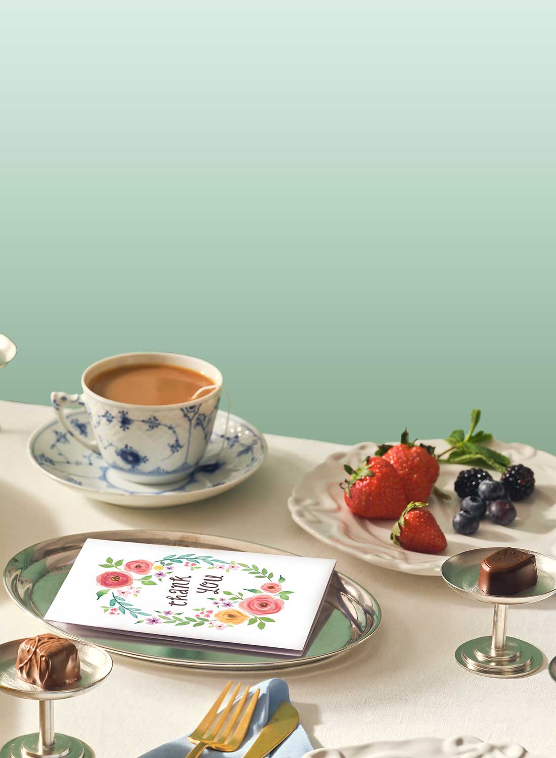 Tea cup with saucer, plate of strawberries and berries, and a 'Thank You' card on a table with a light green background.