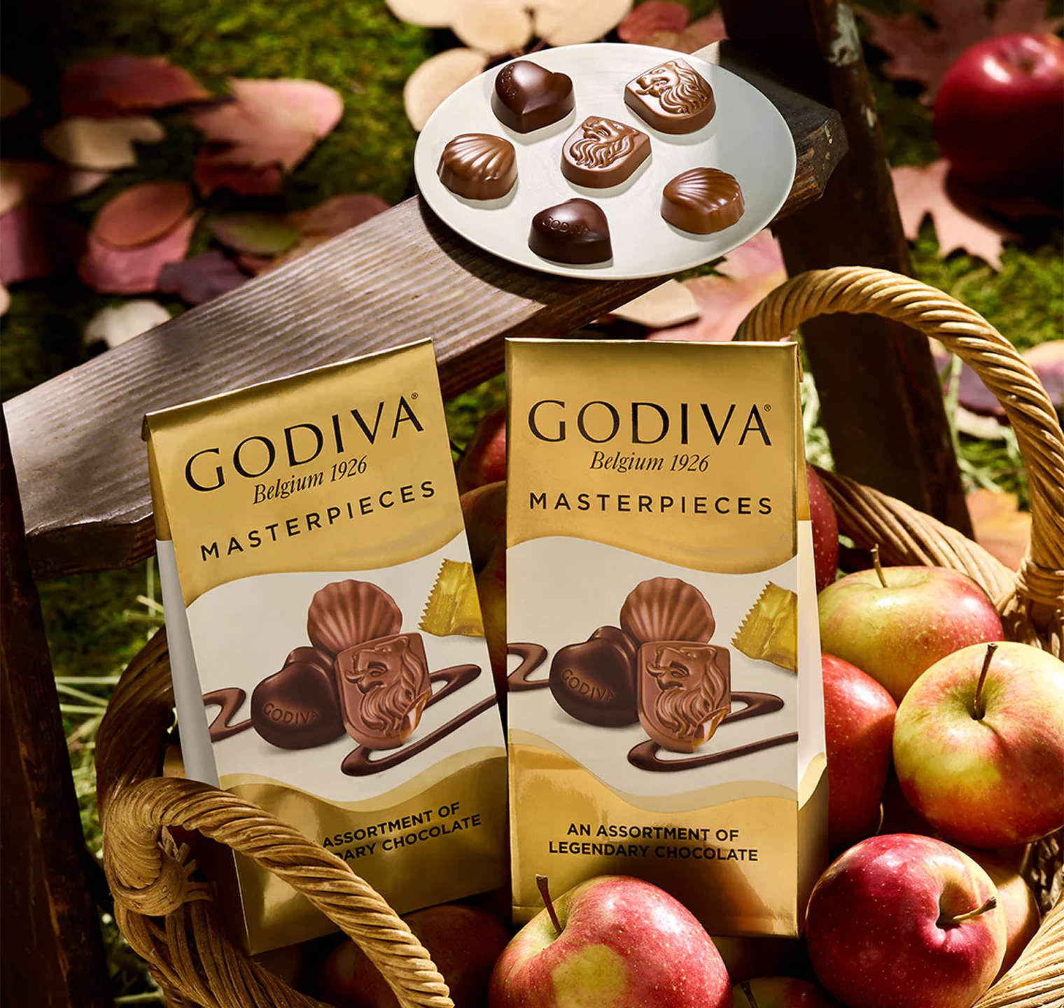 Two Godiva Masterpieces chocolate boxes with a plate of chocolates and apples in the background.